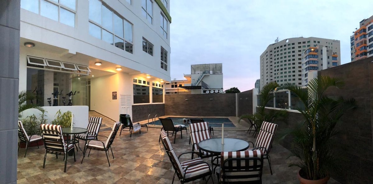 An outdoor area is shown with various seating arrangements, including striped lounge chairs and a circular dining table. A pool can be seen in the background, and the setting is surrounded by walls with greenery, enhancing the spacious appearance of the space.
