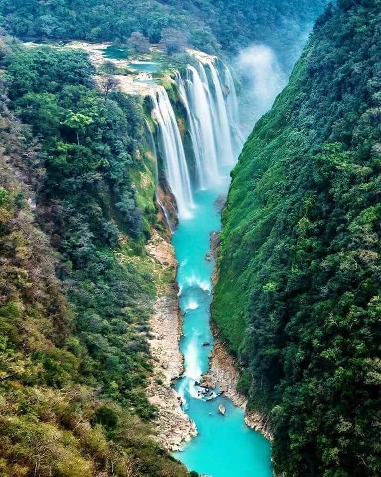 A breathtaking view of a cascading waterfall is captured, surrounded by lush green cliffs and a vibrant blue river below. The mist from the falls rises into the air, creating a serene atmosphere within this natural setting.
