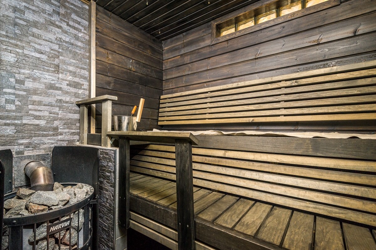 A wooden sauna interior features bench seating along the walls and a stone fireplace with a metal stove. Soft lighting highlights the natural wood grain, creating a serene setting for relaxation and rejuvenation.