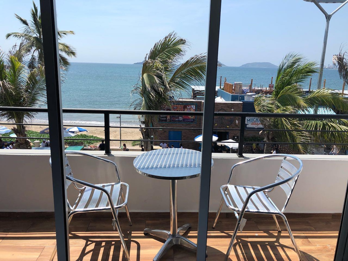 A balcony is visible, featuring a small round table accompanied by two silver chairs. The view showcases the beach and ocean, framed by swaying palm trees. The scene captures a serene coastal atmosphere, enhancing the overall connection to the surrounding environment.