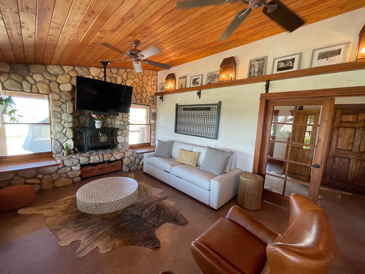 A cozy living room showcases a stone fireplace with a wood mantle, accompanied by comfortable seating options. Light streams in through multiple windows, providing a view of the outside. Warm wood tones on the ceiling and walls create an inviting ambiance.