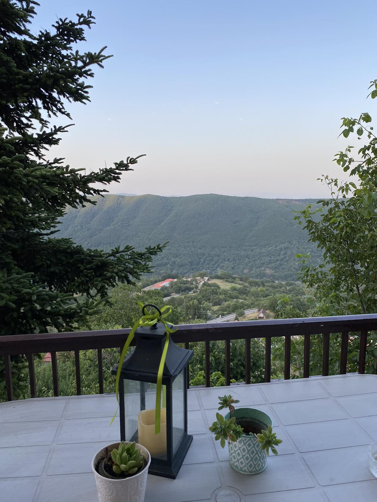 A serene view of rolling green hills is framed by a wooden railing. A lantern sits on a table alongside potted plants, offering a welcoming touch to the outdoor space. The soft evening light enhances the peaceful atmosphere of the surroundings.