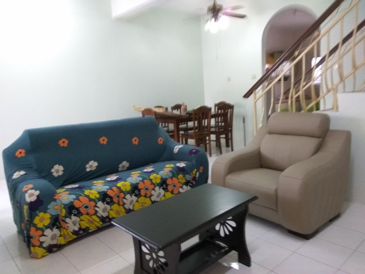 A living area features a floral-patterned couch alongside a comfortable armchair. A dark coffee table occupies the center, while a dining area with wooden chairs and a table is visible in the background. Light-colored walls create a spacious and inviting feel.