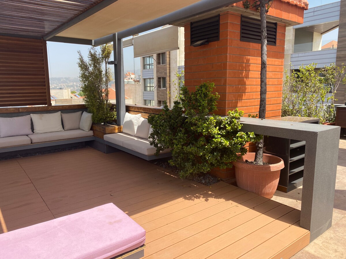 A spacious terrace features comfortable seating areas, including a long bench with soft cushions and lush potted plants. A sleek outdoor kitchen space is visible, complemented by a wooden deck that enhances the area’s inviting atmosphere.