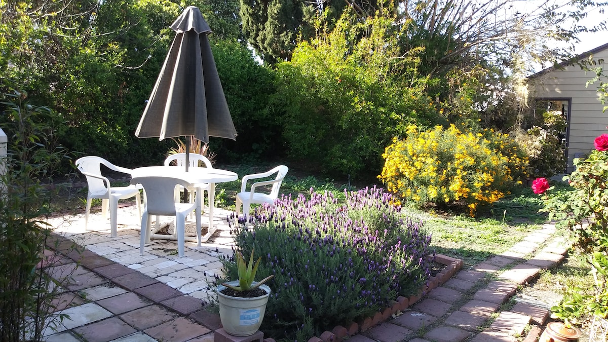 A shaded patio area is equipped with a large umbrella and four white chairs. Lush greenery surrounds a vibrant yellow flowering bush and purple lavender plants. A pathway made of pavers leads through the garden, providing a serene setting for relaxation.