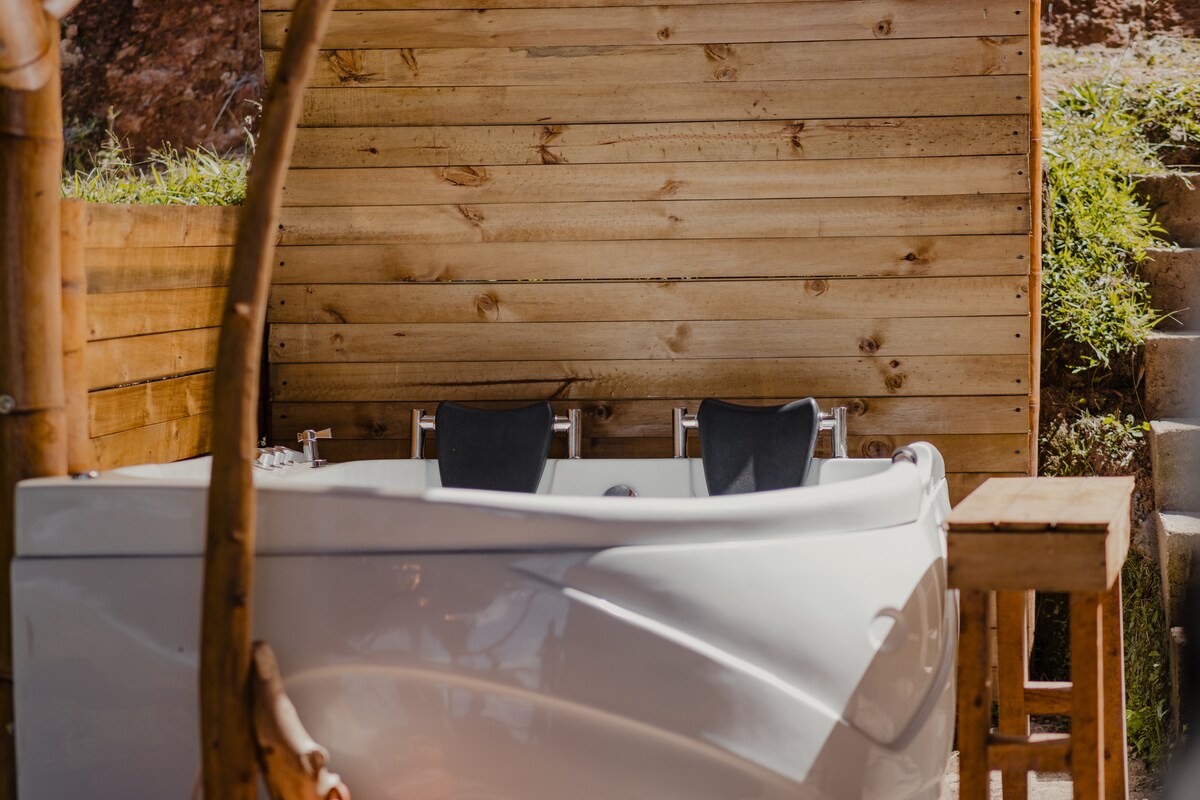 A private outdoor jacuzzi is set against a wooden backdrop, providing a serene space for relaxation. Two comfortable chairs are positioned nearby, complemented by a wooden table that enhances the rustic charm of the area surrounded by greenery.