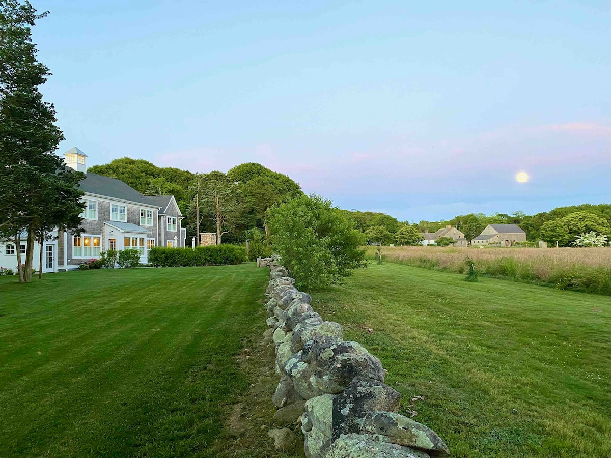 A serene landscape is depicted, featuring a well-maintained grassy area with a low stone wall. The home stands to one side, framed by trees, while a tranquil field extends towards the horizon under a soft twilight sky, highlighting a rising moon.