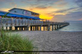 An expansive sandy beach leads to a wooden pier extending over calm waters. The structure features a restaurant with blue awnings, softly illuminated by the warm colors of a sunset. Gentle waves lap at the shore, creating a serene coastal scene.