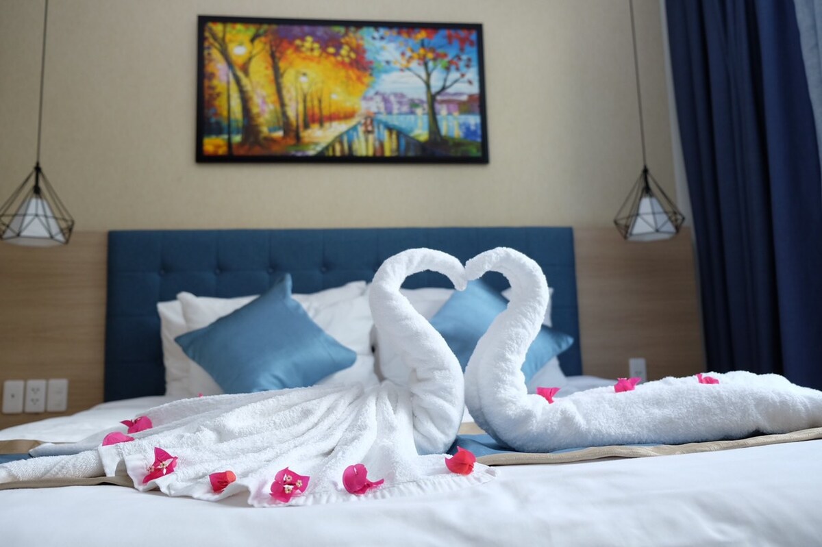 A spacious bed is adorned with decorative towel swans, crafted into a heart shape. Bright blue pillows and scattered pink petals complement the bedding. A colorful painting featuring a serene landscape hangs above the bed, enhancing the room's calming ambiance.