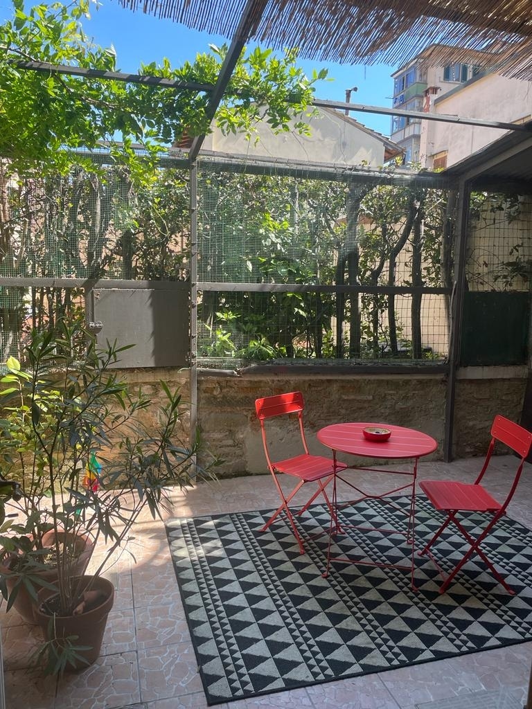 A private garden space features a patterned rug beneath a small, round red table surrounded by two matching chairs. Lush greenery is visible along the perimeter, providing a sense of privacy. Sunlight filters through the overhead structure, creating a bright and inviting area.
