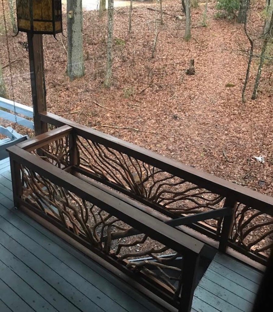 A wooden railing with an intricate tree branch design overlooks a serene forest landscape, where the ground is covered with fallen leaves. The view from the porch showcases a peaceful outdoor setting, framed by tall trees, providing a sense of tranquility.