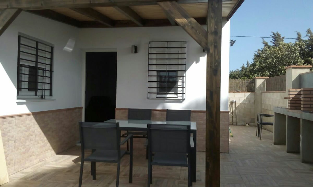 A covered terrace area features a glass-top dining table complemented by four chairs. Sunlight filters through the structure, highlighting the white walls and stone accents. A doorway leads to the interior, with a glimpse of the outdoor area beyond.