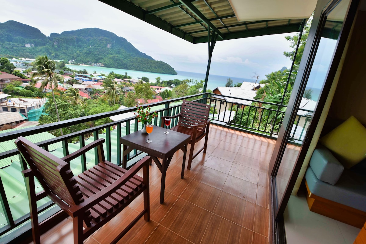 A private balcony showcases a scenic view of the coastline and surrounding hills. Two wooden chairs are seated at a small table, complemented by a bright orange drink. Large glass doors offer access to the interior space, enhancing the connection with the outdoors.