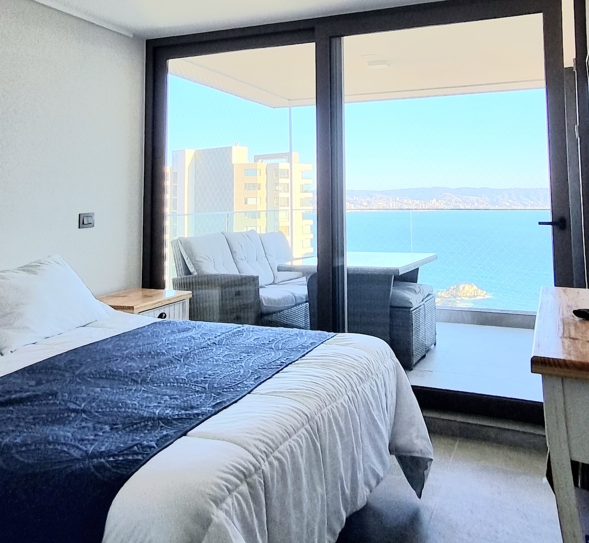 A spacious bedroom is featured, with a large bed covered by a white quilt. Floor-to-ceiling glass doors provide access to a private balcony with views of the ocean. A cozy seating area with two chairs is visible, ideal for relaxation.