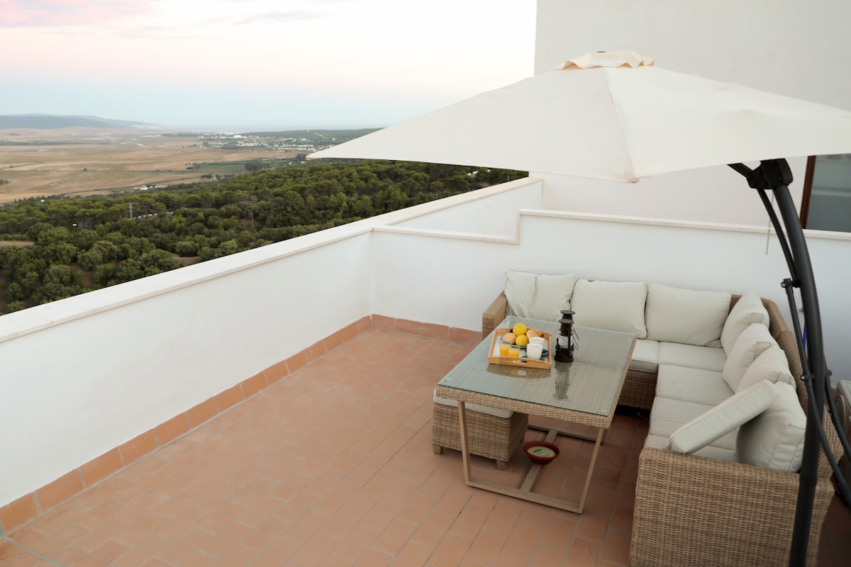 A spacious private terrace featuring a sectional sofa with neutral cushions is complemented by a glass-top table set with a fruit bowl and beverage. An umbrella provides shade, while the expansive view of the surrounding landscape is visible in the background.
