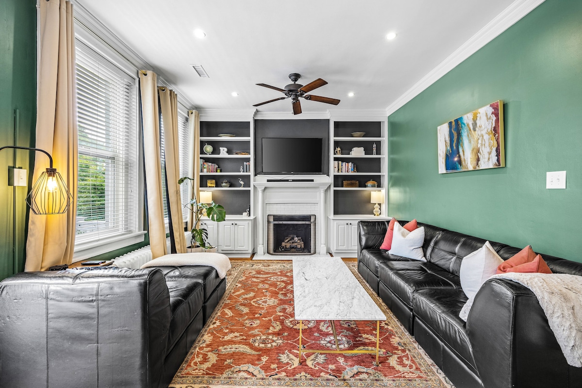The living area showcases a marble-topped coffee table centered between two black leather sofas. A ceiling fan and recessed lighting provide illumination. Shelving units display decorative items, while a mounted TV is framed by a white mantel. Natural light filters through the windows adorned with light curtains.
