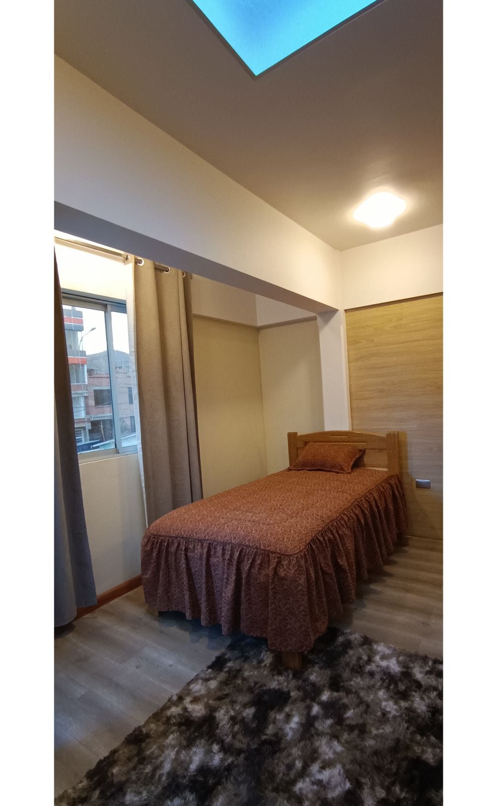 A cozy sleeping area is equipped with a single bed, adorned with a patterned bedspread. Natural light enters through a nearby window, which is framed by curtains. The room features warm-toned walls and wooden accents, creating a welcoming space.