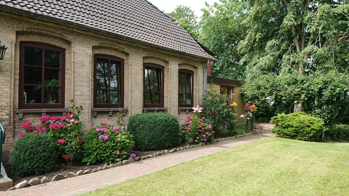 A charming exterior view of a house features large windows framed with dark wood. Flowering shrubs in pink and purple bloom along the pathway, leading to a well-maintained lawn surrounded by greenery, enhancing the outdoor space.