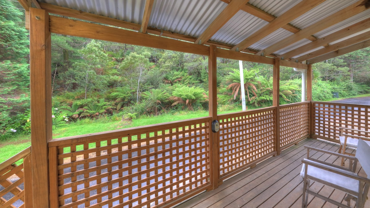 A covered wooden deck features a lattice design, offering views of a lush green landscape. The surrounding foliage includes ferns, enhancing the natural setting. Two chairs are positioned for seating, providing a space to relax outdoors.