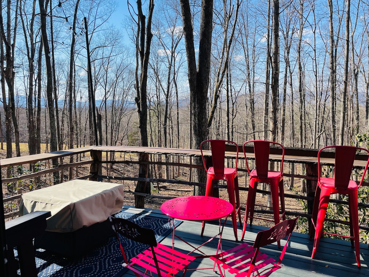 A spacious outdoor deck is surrounded by tall, bare trees, offering a clear view of the landscape. Bright red metal chairs are arranged around a matching table, providing a comfortable spot to enjoy the natural surroundings. A large, covered area is visible beneath a protective cover.