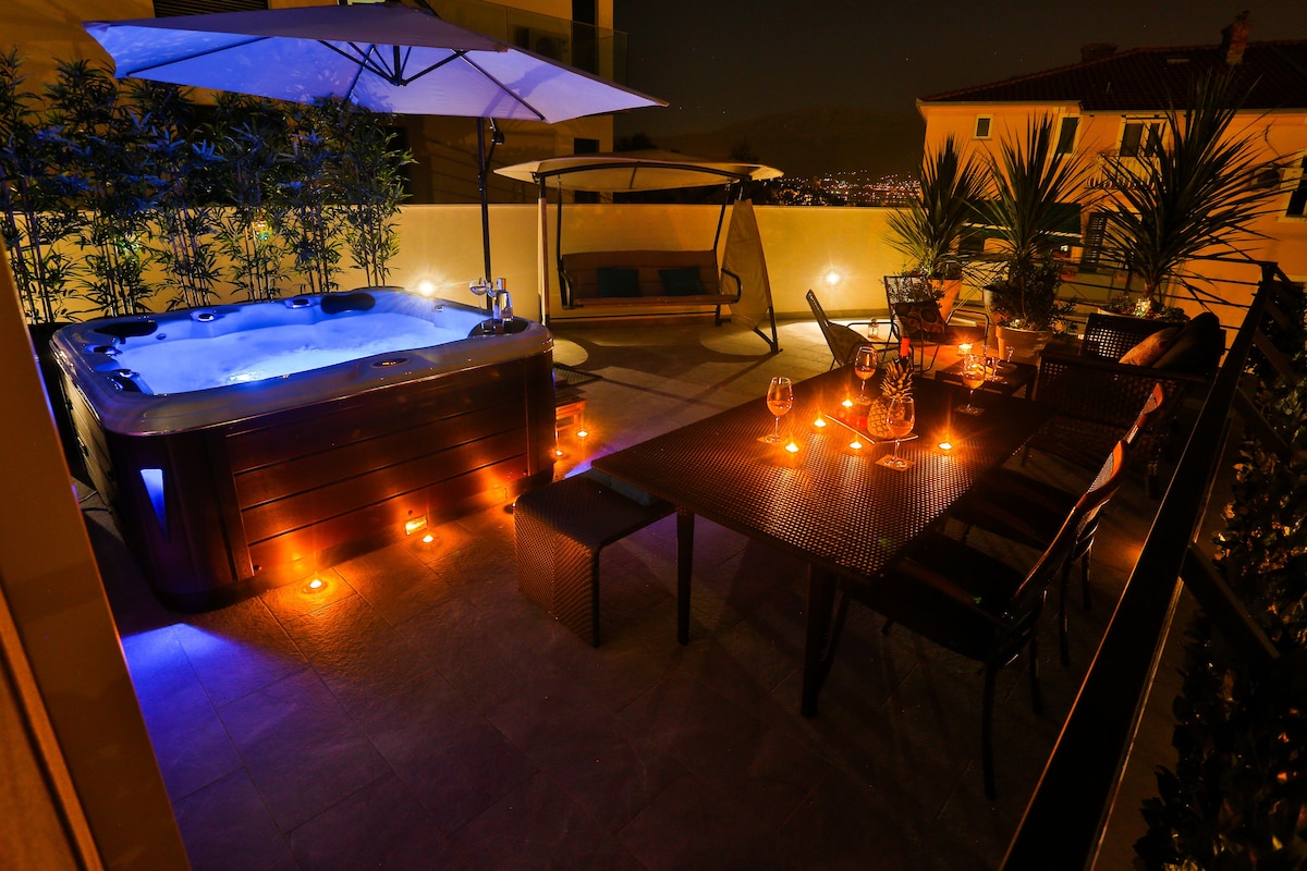 An outdoor terrace features a jacuzzi with ambient lighting, surrounded by plants. A dining area with a table and chairs is set for multiple guests, complemented by a seating area with a swing. Soft glow from candles enhances the relaxing atmosphere.