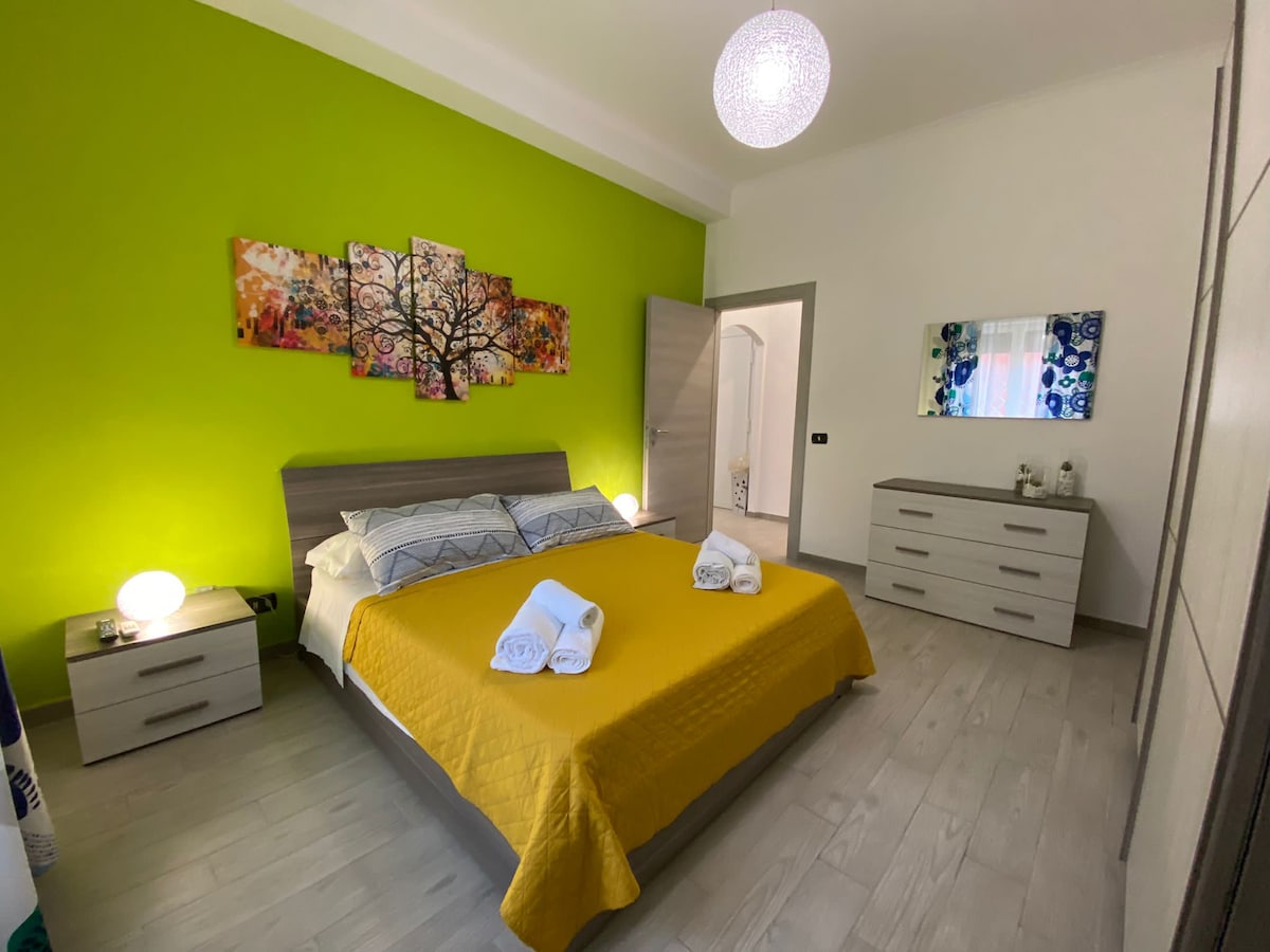 A modern bedroom features a spacious bed with a yellow blanket, complemented by two bedside lamps. A vibrant green accent wall enhances the room. Artwork depicting trees adds a splash of color above the bed. Neutral tones in the furniture create a balanced atmosphere.