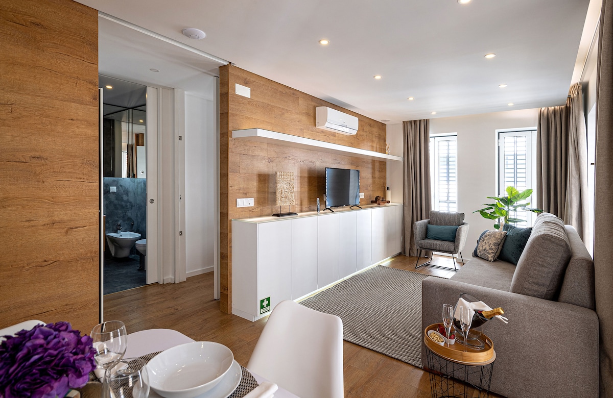 A bright living area features a comfortable gray sofa and a small coffee table, with a television mounted on a sleek white unit. The space is enhanced by wooden paneling and natural light coming through large windows. A dining table is set nearby, ready for meals.