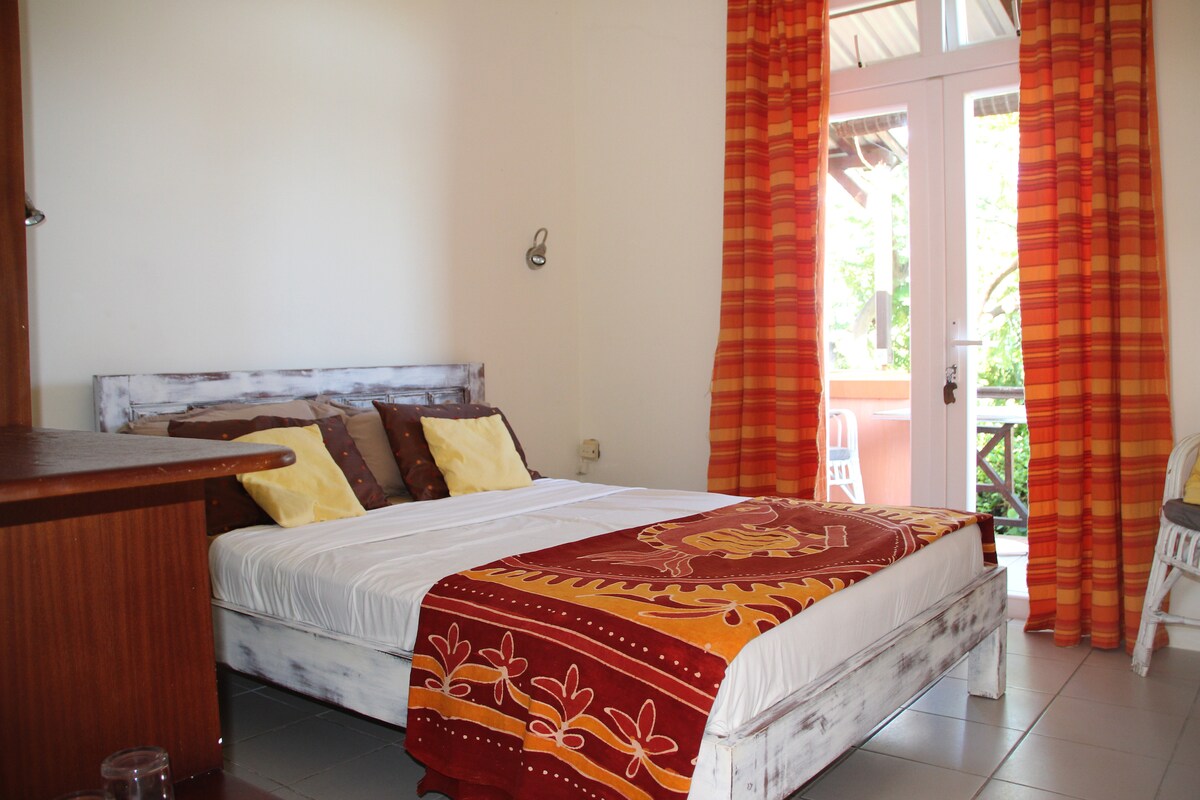 A small but cozy room is showcased, featuring a spacious bed adorned with decorative pillows and a vibrant blanket. Large glass doors open to a private balcony, inviting natural light, while bright orange curtains frame the scene.