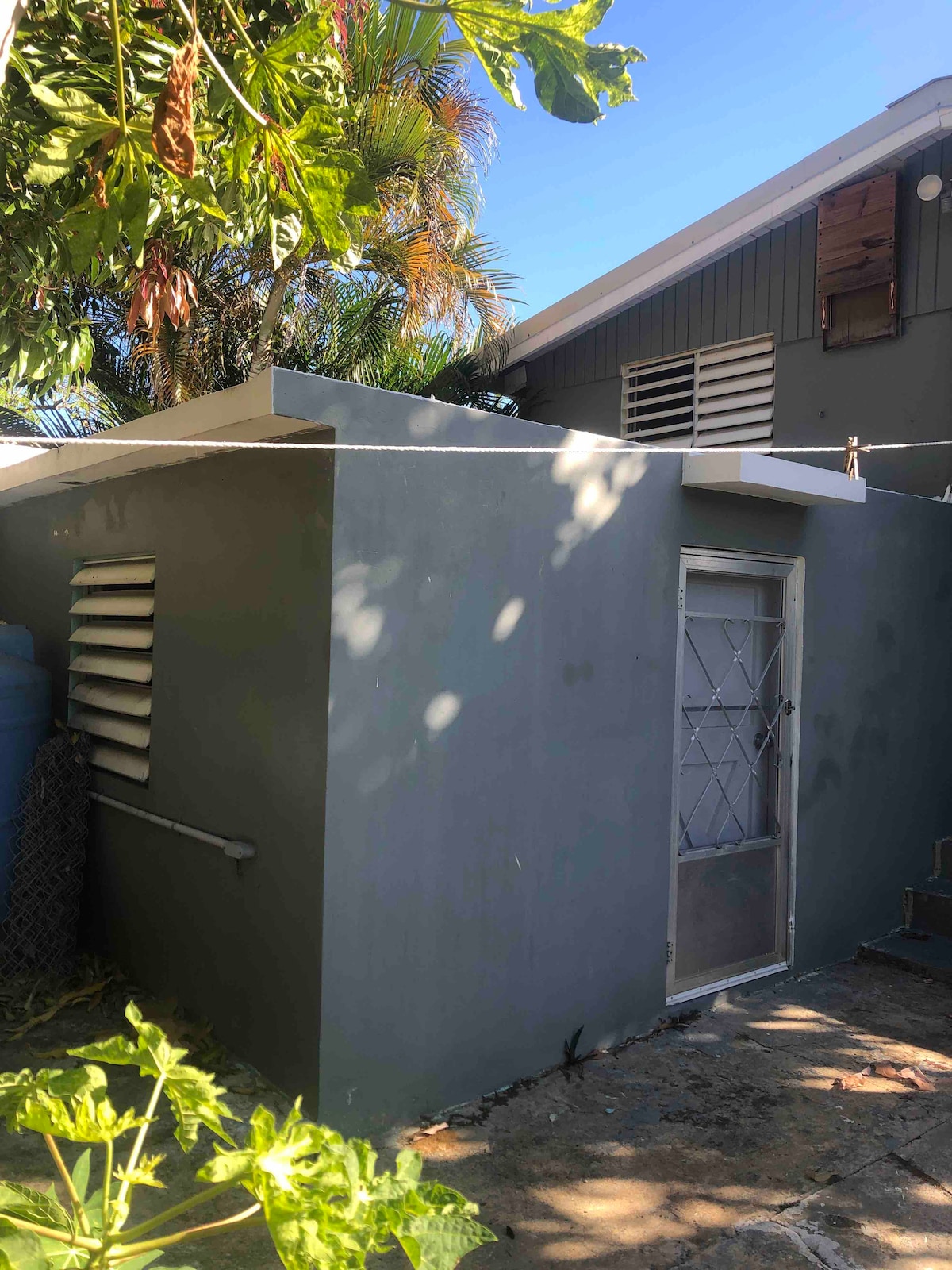 A small gray structure is visible, partially shaded by surrounding trees. The exterior features a simple door with a diamond-pattern grate. Sunlight casts soft shadows on the weathered stone floor, providing a glimpse of the tropical landscaping present around the building.