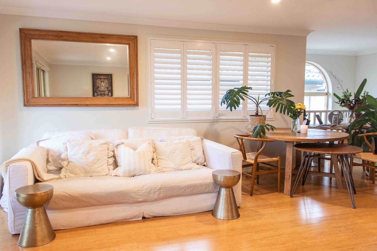 A bright and open living area is showcased with a white sofa adorned with decorative pillows. A round wooden dining table is present, surrounded by light-colored chairs. Natural light filters through the window with plantation shutters, enhancing the relaxed atmosphere.