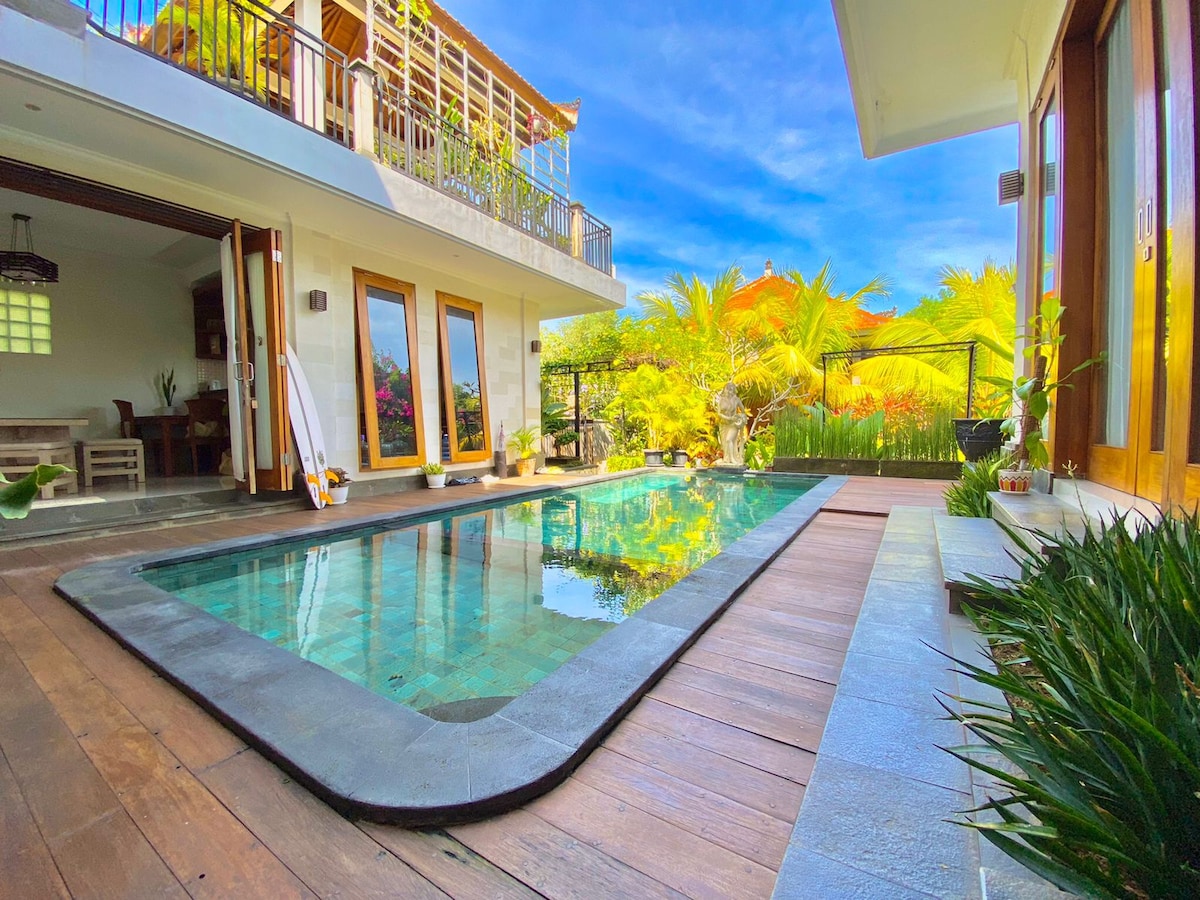 The private pool, framed by wooden decking, reflects the clear blue sky. Lush greenery surrounds the area, enhancing the natural ambiance. Open doors lead to the inviting indoor spaces, blending the outdoor and indoor environments seamlessly.