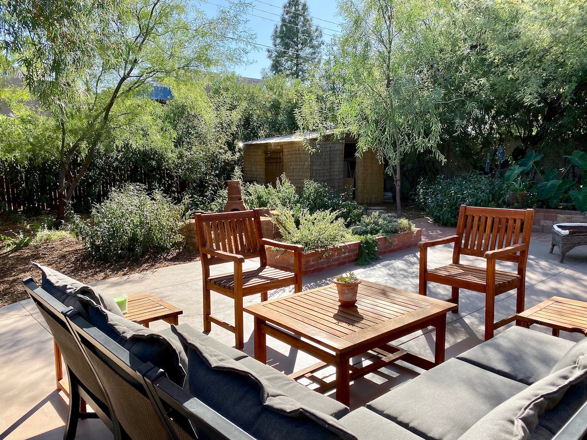 An outdoor seating area features wooden chairs and a coffee table arranged on a paved patio. Lush greenery surrounds the space, including various plants and trees. A rustic structure can be seen in the background, providing a cozy element to the garden atmosphere.