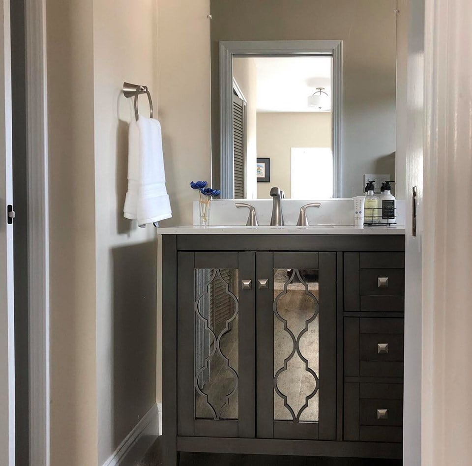 A modern bathroom features a stylish vanity with mirrored cabinet doors and dual sinks. Soft natural light filters through a doorway in the background, illuminating the space. A towel is neatly hung on a chrome rack, while personal care items are organized on the counter.
