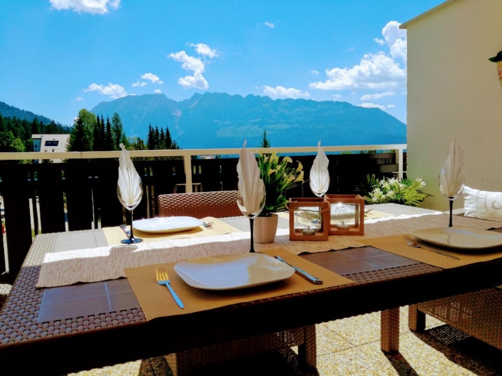 A dining table is set with plates, cutlery, and decorative napkins, overlooking a stunning mountainous landscape. Sunlight brightens the space, creating an inviting atmosphere for outdoor meals. Lush greenery is visible in the background, enhancing the serene environment.