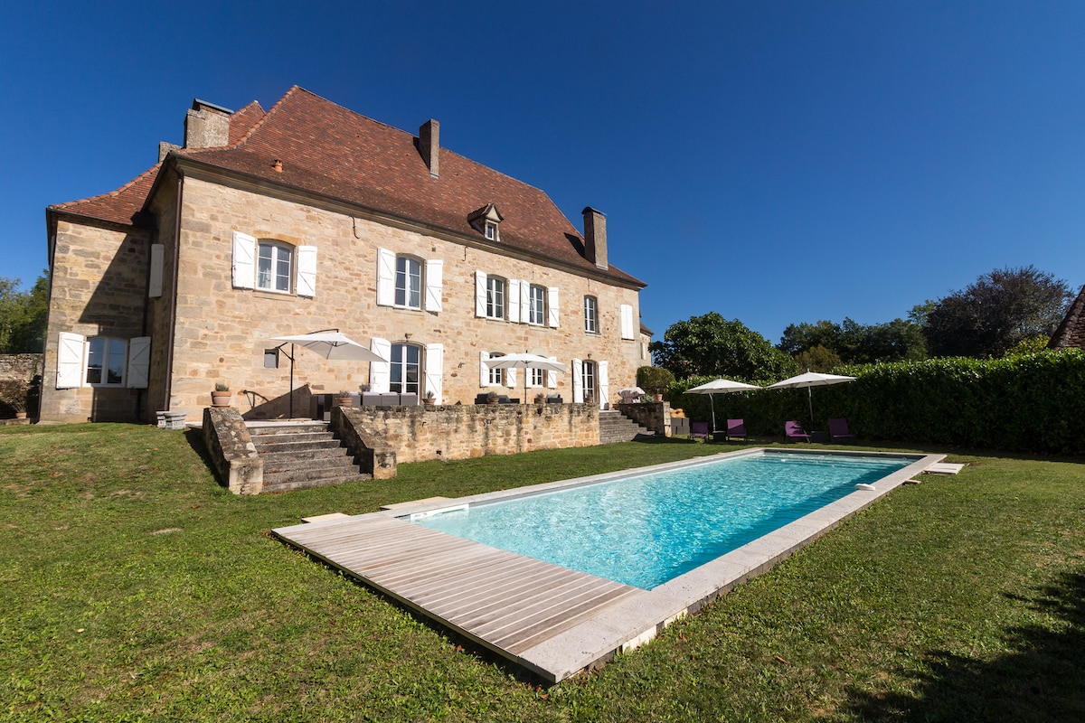 The exterior of a spacious stone manor house is depicted, showcasing a well-maintained garden with a large swimming pool in the foreground. Sun loungers are arranged alongside the water, while the clear blue sky and vibrant greenery create an inviting outdoor retreat.