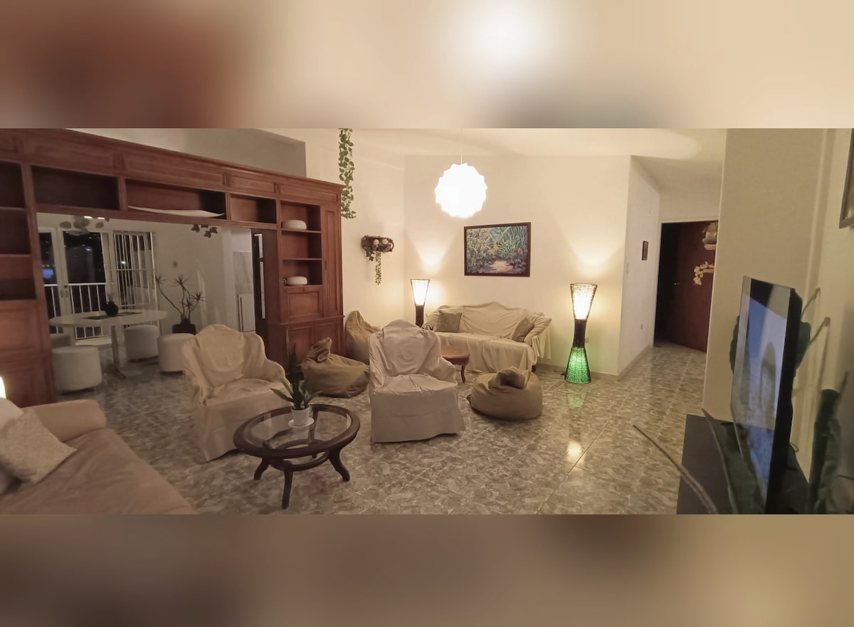 A spacious living area features comfortable seating arranged around a coffee table. Soft lighting from floor lamps and a central fixture creates a relaxed ambiance. A television is positioned on a shelf, with artwork displayed on the wall, complemented by decorative plants.