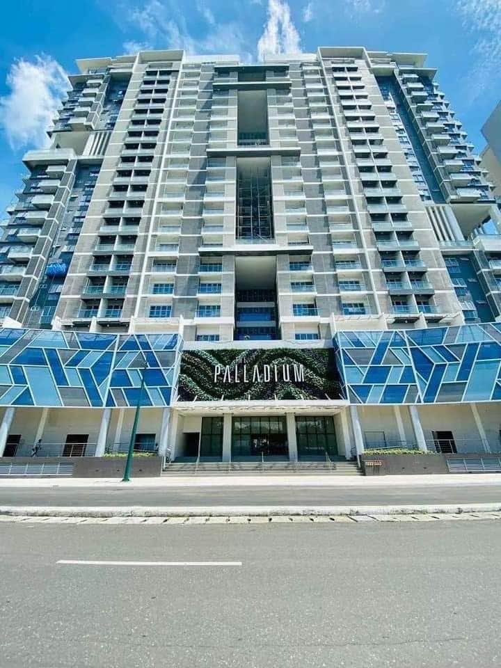 The modern exterior of The Palladium is showcased, featuring a striking façade with geometric glass panels. The building's entrance is highlighted by the name 'Palladium' prominently displayed, while a wide pathway leads up to the entrance, set against a clear blue sky.