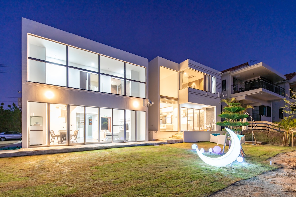 A modern private villa is showcased during twilight, featuring expansive glass walls that reveal interior spaces. An illuminated artistic feature is situated in the well-maintained garden, complementing the sleek exterior design.
