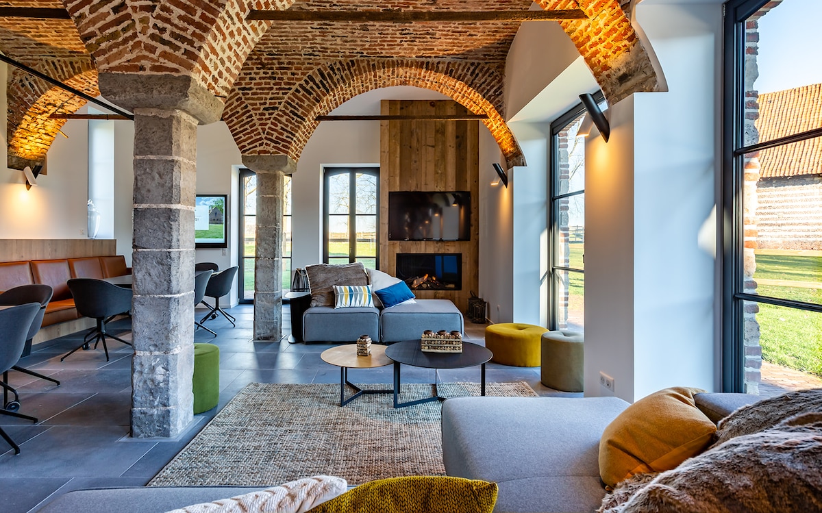 A spacious living area features exposed brick arches and natural stone columns. Soft seating is arranged around low coffee tables, with large windows providing views of the surrounding garden. Subtle lighting highlights the architectural details, creating a comfortable and inviting environment.