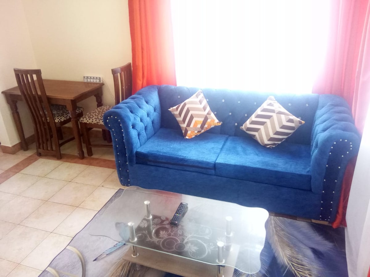 A living area features a blue tufted sofa adorned with decorative pillows, offering a comfortable seating option. A glass coffee table sits in front, and a small wooden dining table with chairs is positioned against the wall. Bright curtains allow natural light to enter the space.