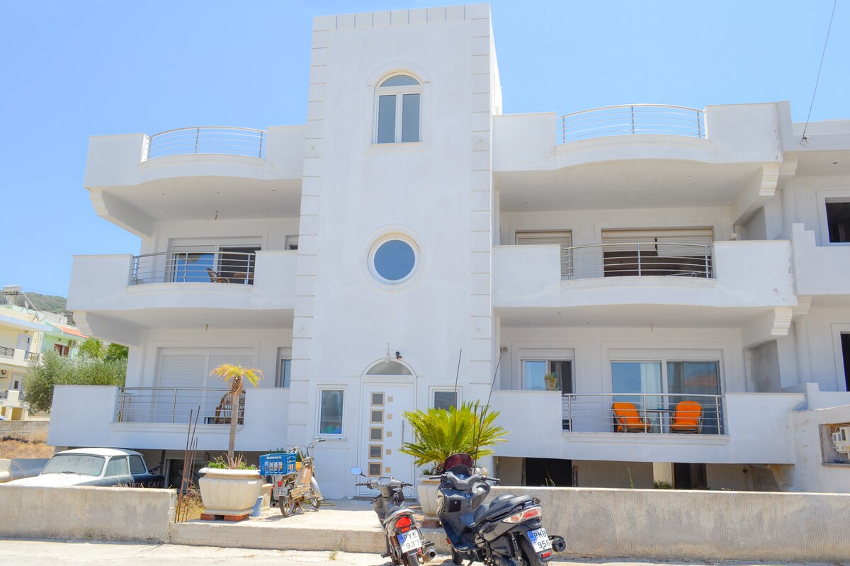 A modern, multi-story building features smooth white exterior walls. Balconies with metal railings project from the facade, offering views of the surroundings. A round window is centered on the ground floor, with two vibrant orange chairs set on a balcony above. Parking spaces for vehicles are visible in front.