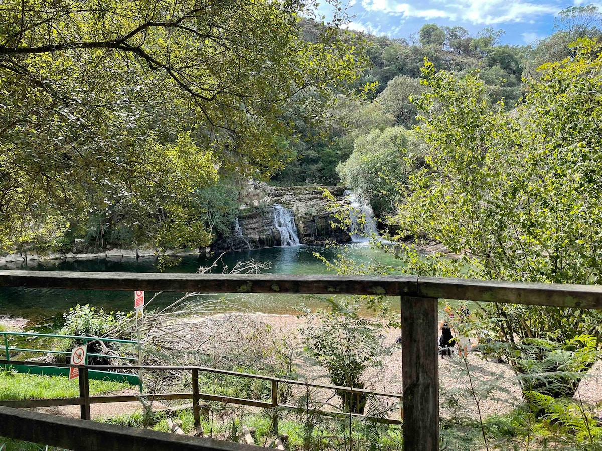 A tranquil scene features a cascading waterfall surrounded by lush greenery. The water flows gracefully into a serene pond, while trees provide a natural frame. A wooden railing is visible in the foreground, offering a vantage point to enjoy the scenery.