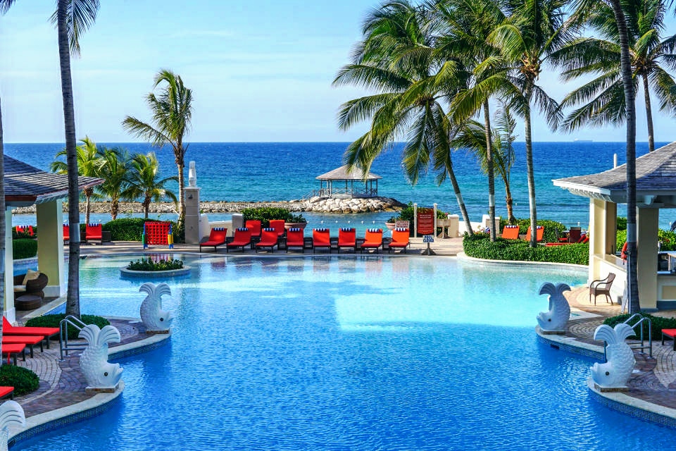 A large swimming pool features vibrant red lounge chairs arranged around it, with palm trees providing shade. The ocean can be seen in the distance, complementing the tranquil atmosphere of the resort. A gazebo is visible along the beachfront, adding to the scenic view.