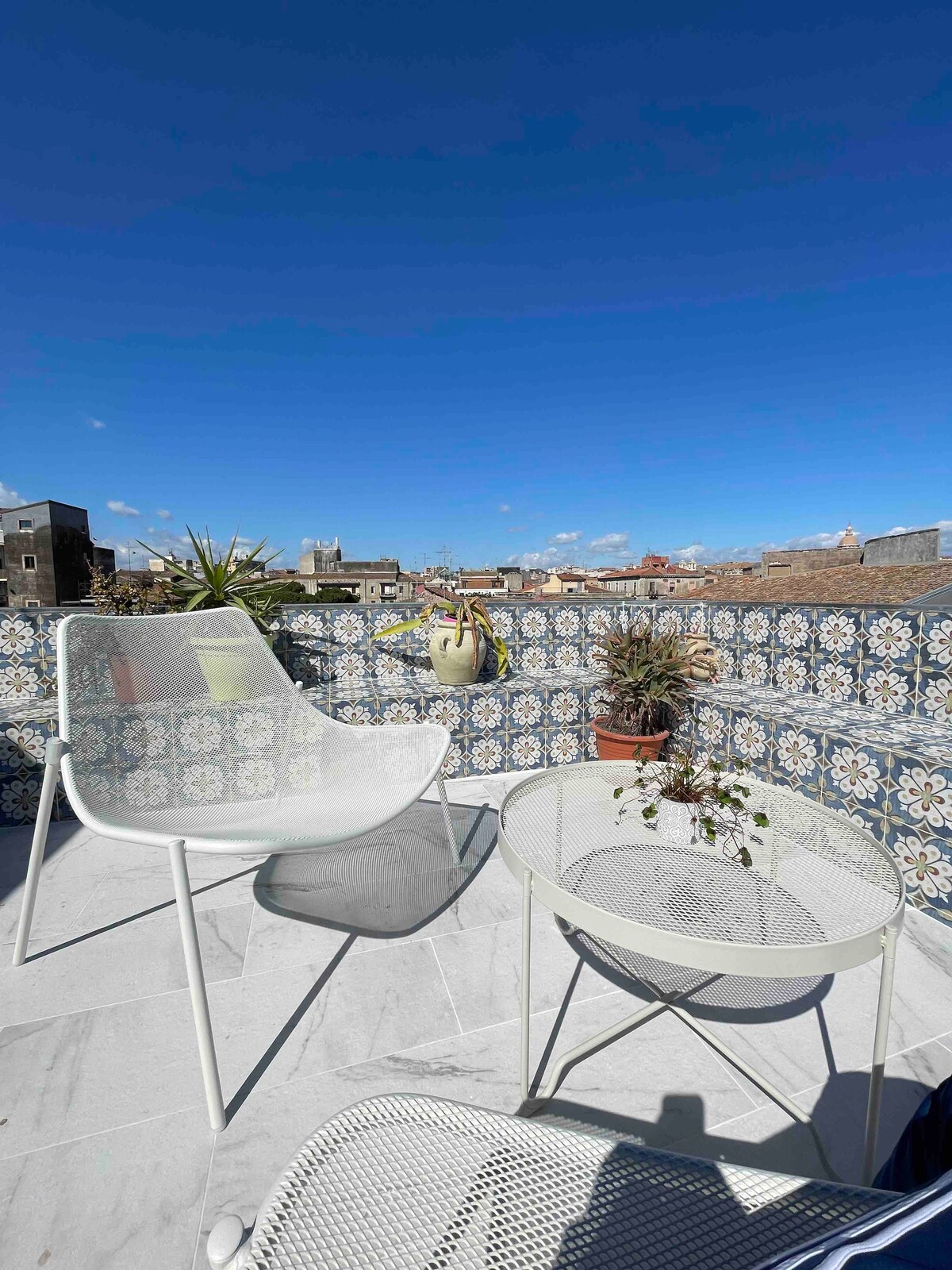 A private terrace is presented, adorned with patterned tiles and potted plants. Two modern white chairs and a round table create a cozy seating area. A clear blue sky extends overhead, providing a serene outdoor space with views of the surrounding rooftops.