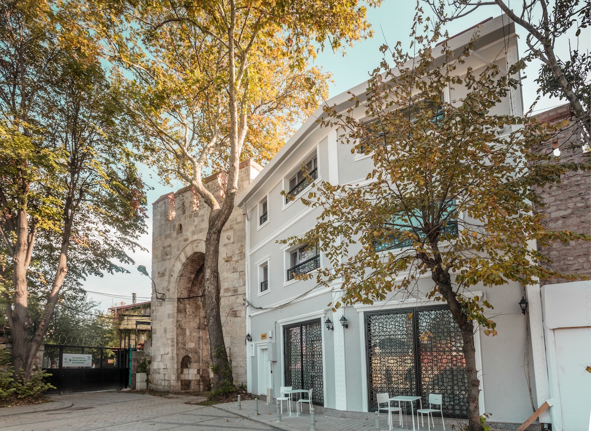 A modern building with white exterior walls stands beside an ancient stone structure, surrounded by large trees. Outdoor seating is positioned in front, featuring several white chairs. Warm sunlight bathes the scene, enhancing the serene atmosphere of the tranquil courtyard.