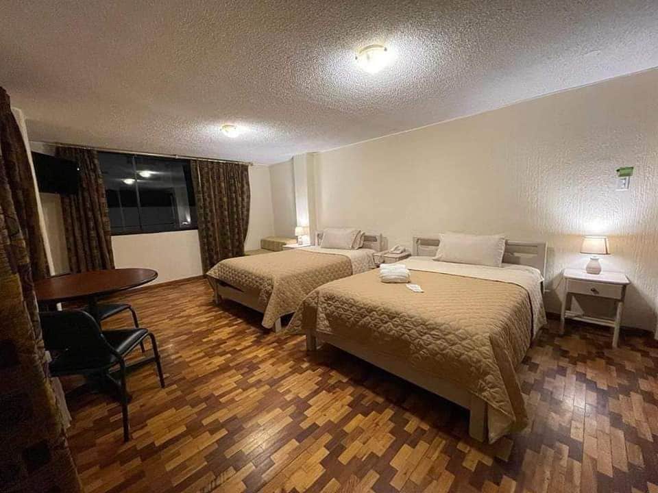 A well-lit room features two spacious beds with neutral-toned bedding. A small table and chairs are positioned near a window dressed with patterned curtains. The warm wooden flooring complements the subtle decor, creating a comfortable space for relaxation.