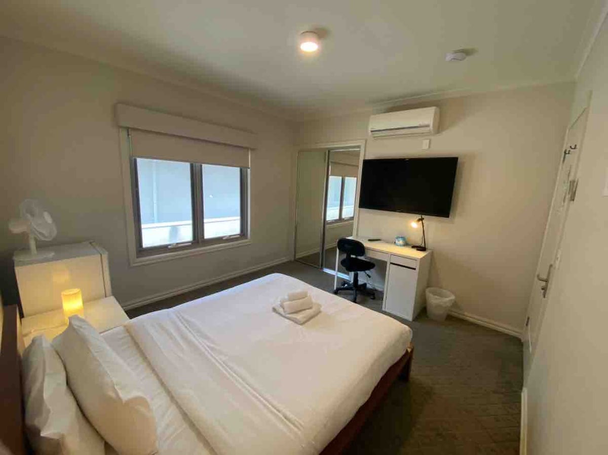 A well-lit bedroom features a spacious bed with fresh linen. A desk and chair provide a workspace, complemented by a wall-mounted TV. Large windows allow natural light, while air-conditioning is available for comfort. A wardrobe and a small waste bin are also present in the room.