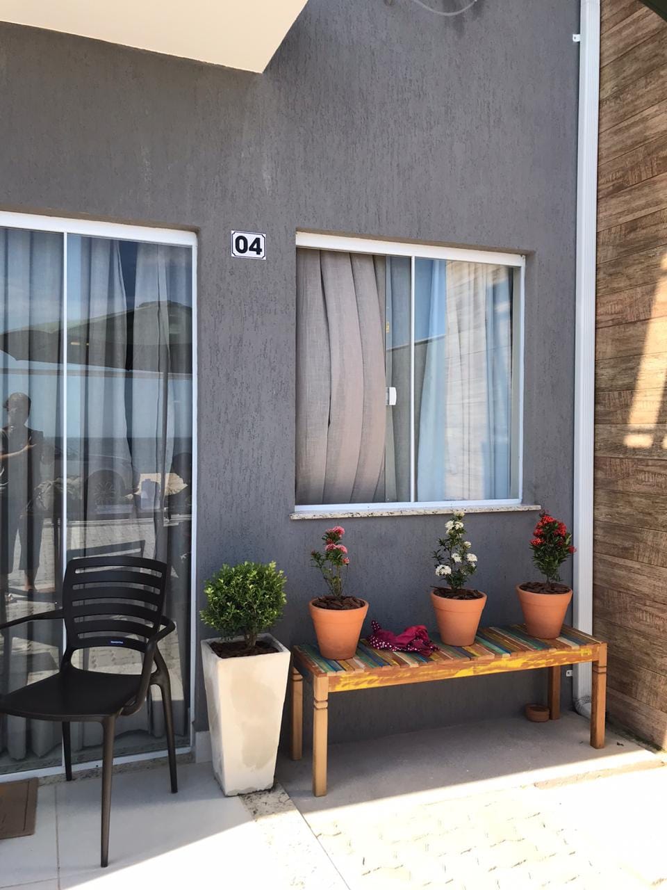 A charming exterior scene features a small bench adorned with potted plants, set against a neutral-colored wall. A window is flanked by sheer curtains, providing a hint of privacy. A chair is positioned nearby, enhancing the inviting entrance.
