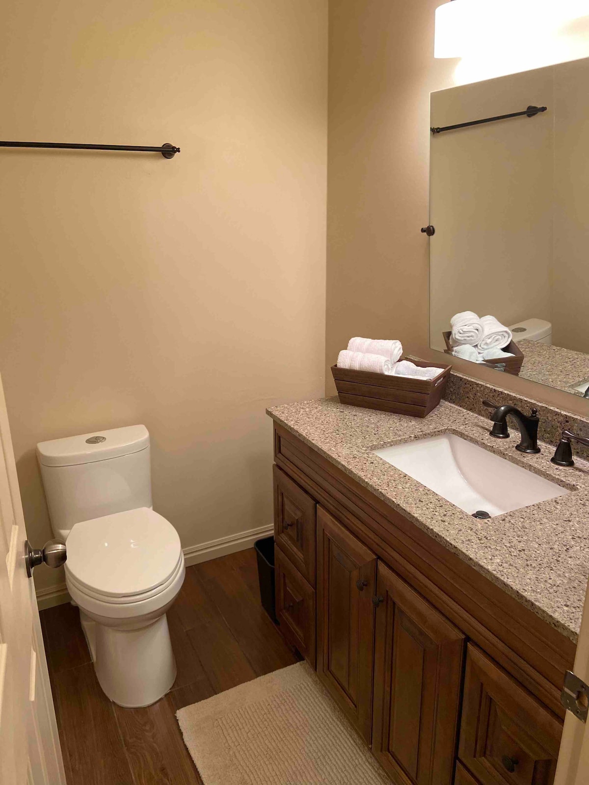 A well-appointed bathroom features a modern toilet and a granite countertop with an under-mount sink. A large mirror reflects soft lighting, while neatly arranged towels are presented in a basket. The floor is adorned with a small rug, complementing the wooden cabinetry.