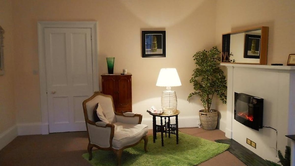 A cozy sitting area features a light-colored armchair beside a small table, accompanied by a lamp. A potted plant adds a touch of greenery, while a wall-mounted mirror reflects light in the room. Soft carpet covers the floor, enhancing the inviting feel.
